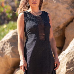 Black sleeveless hi-low ethnic dress in handwoven rustic cotton with round neckline, intentional distressed frayed detail at chest and dramatic asymmetric skirt short at front and long at back with raw frayed hem, worn against rocky coastal background - Raja Pura artisan boho dress - portrait front view