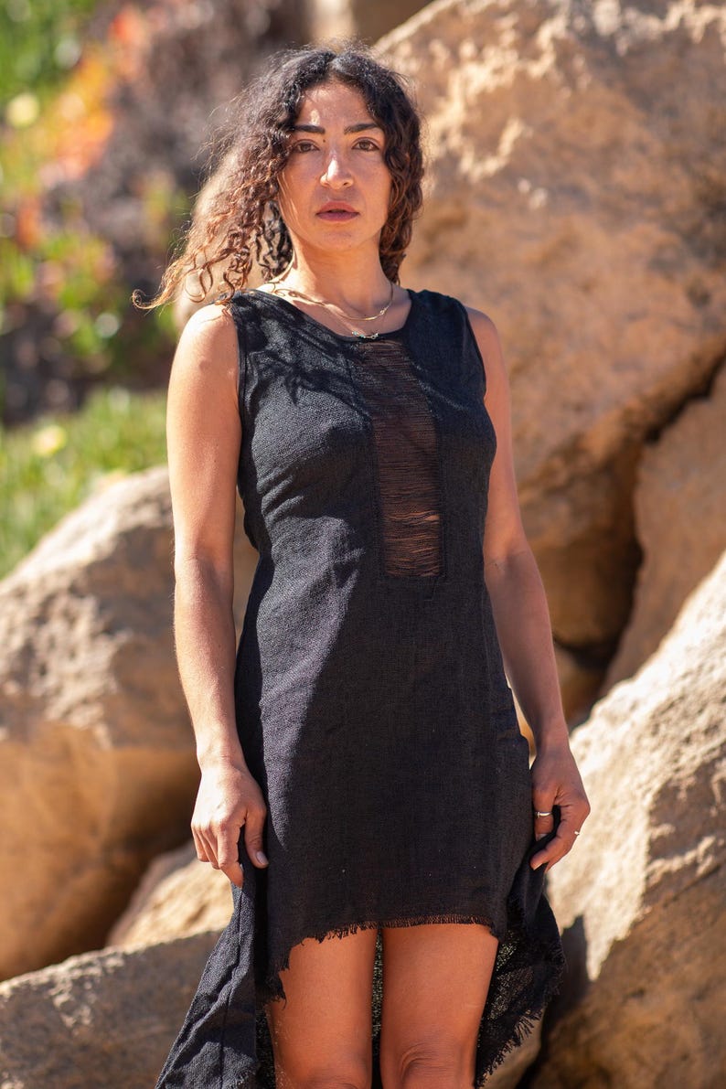 Black sleeveless hi-low ethnic dress in handwoven rustic cotton with round neckline, intentional distressed frayed detail at chest and dramatic asymmetric skirt short at front and long at back with raw frayed hem, worn against rocky coastal background - Raja Pura artisan boho dress - portrait front view