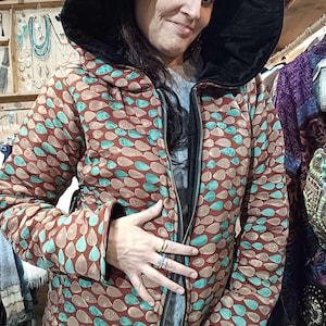 Peut inclure: Veste à capuche avec un motif de gouttes d'eau marron, turquoise et beige. La veste est dotée d'une capuche doublée de velours noir et d'une fermeture éclair intégrale. Vêtement de la collection.