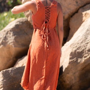 Terracotta sleeveless hi-low ethnic dress in handwoven rustic cotton with round neckline, intentional distressed frayed detail at chest and dramatic asymmetric skirt short at front and long at back with raw frayed hem, worn against rocky coastal background - Raja Pura artisan boho dress - full body back view