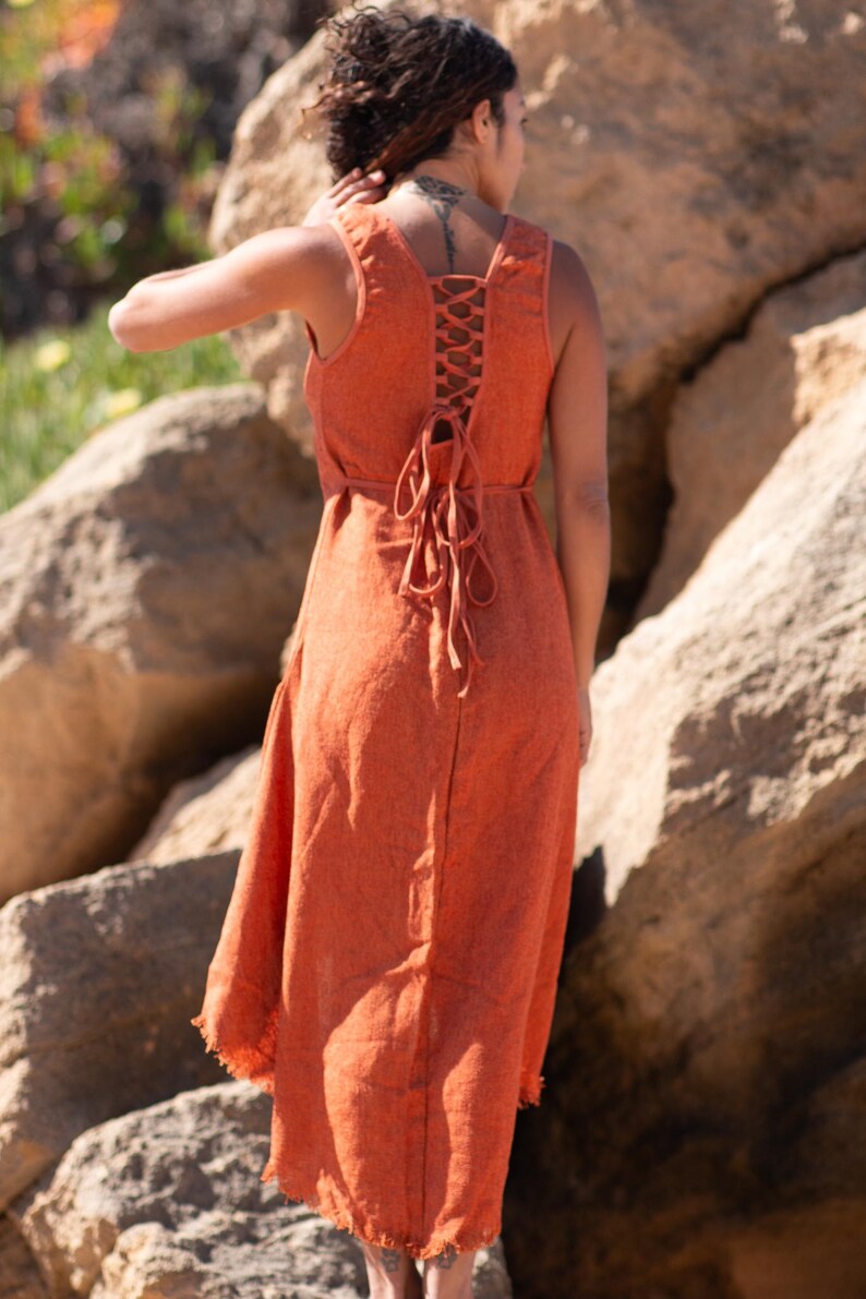 Terracotta sleeveless hi-low ethnic dress in handwoven rustic cotton with round neckline, intentional distressed frayed detail at chest and dramatic asymmetric skirt short at front and long at back with raw frayed hem, worn against rocky coastal background - Raja Pura artisan boho dress - full body back view