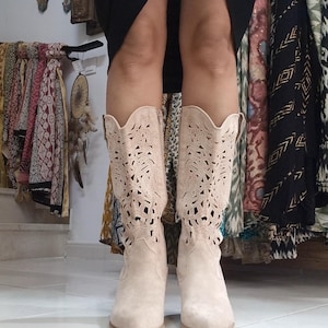 Peut inclure: Une paire de bottes de cowboy en daim beige clair. Les bottes présentent un motif ajouré décoratif sur la tige et un bout pointu. Les bottes sont hautes et ont un petit talon.