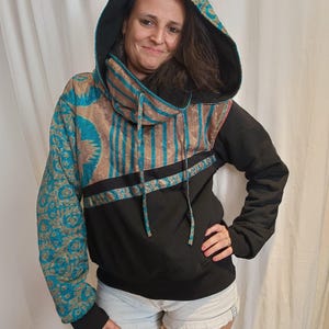 Peut inclure: Sweat à capuche noir avec capuche et manche à motifs turquoise et or. Le sweat à capuche a un corps et des poignets noirs, avec un motif turquoise et or sur la capuche et le haut de la manche. La capuche a un cordon de serrage.