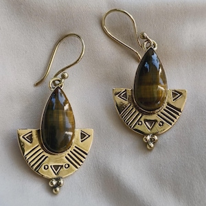 Peut inclure: Une paire de boucles d'oreilles pendantes dorées avec une pierre œil-de-tigre en forme de goutte, surmontant une plaque métallique semi-circulaire gravée de lignes géométriques, de triangles et d'un petit groupe de trois perles.