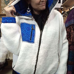 Peut inclure: Veste en polaire blanche à capuche, avec un tissu bleu à motifs sur la doublure de la capuche, les poignets et la poche. La veste a une fermeture éclair sur toute la longueur et une petite poche avec une fermeture éclair. La capuche est bordée d'un tissu à motifs.