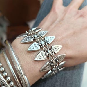 Peut inclure: Un bracelet en argent avec un design géométrique composé d'éléments stylisés en forme de flèche et de détails complexes. Le bracelet est relié par de petites chaînes et orné de petits accents ronds. Plusieurs autres bracelets en argent sont également visibles.