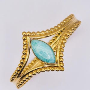 Peut inclure: Un bracelet manchette doré orné d'une pierre centrale bleu clair en forme de marquise avec des marbrures blanches. La monture métallique présente une texture perlée sur les bords, formant un losange géométrique autour de la pierre.