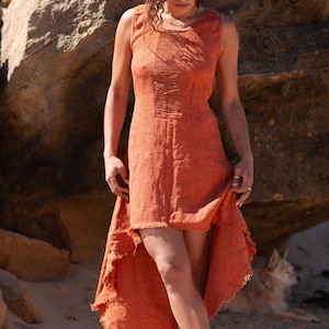 Terracotta sleeveless hi-low ethnic dress in handwoven rustic cotton with round neckline, intentional distressed frayed detail at chest and dramatic asymmetric skirt short at front and long at back with raw frayed hem, worn against rocky coastal background - Raja Pura artisan boho dress - full body front view