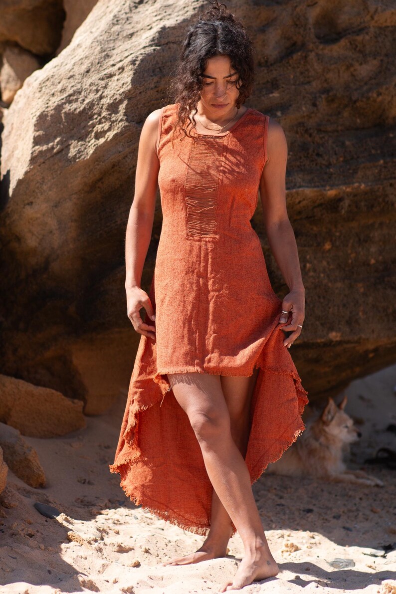Terracotta sleeveless hi-low ethnic dress in handwoven rustic cotton with round neckline, intentional distressed frayed detail at chest and dramatic asymmetric skirt short at front and long at back with raw frayed hem, worn against rocky coastal background - Raja Pura artisan boho dress - full body front view