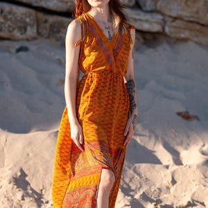Peut inclure: Une robe longue sans manches jaune moutarde avec un décolleté en V, des bretelles à nouer et un imprimé cachemire rouge et orange vif sur l'ourlet et le décolleté. Le vêtement présente une taille ajustée et une jupe longue fendue à l'avant.