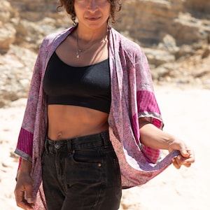 Może przedstawiać: Kobieta w różowo-fioletowym kimono w wzory. Szal ma kwiatowy wzór i jest połączony z czarnym stanikiem sportowym i ciemnymi dżinsami. Tło to piaszczysty, skalisty obszar.