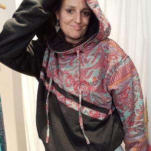 Może przedstawiać: Czarna i wzorzysta bluza z kapturem z wzorem paisley w odcieniach czerwieni, turkusu i pomarańczy. Bluza z kapturem ma czarną dolną część i podszewkę kaptura, ze sznurkami. Rękawy mają wzorzysty mankiet.