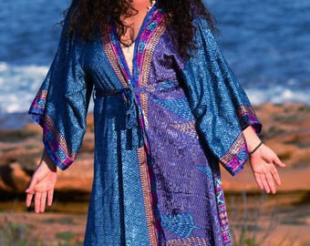 May include: A long, flowing robe in shades of blue and purple, featuring intricate geometric and leaf patterns. The robe has wide sleeves and a tie closure at the waist. Accents of pink and gold trim the edges, creating a bohemian aesthetic.