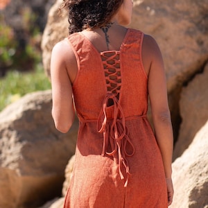 Terracotta sleeveless hi-low ethnic dress in handwoven rustic cotton with round neckline, intentional distressed frayed detail at chest and dramatic asymmetric skirt short at front and long at back with raw frayed hem, worn against rocky coastal background - Raja Pura artisan boho dress - portrait back view