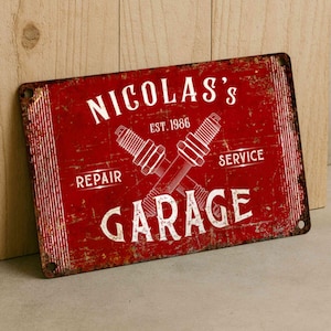 以下が含まれることがあります： 赤色の長方形の金属製サイン。白い文字で「NICOLAS'S GARAGE」、単語「REPAIR」と「SERVICE」、年号「EST. 1986」が記されています。サインには交差したスパークプラグが描かれ、使い古されたヴィンテージの外観をしています。