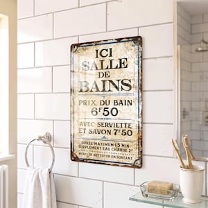 May include: A rectangular metal sign with a distressed, off-white background and blue border. The text reads: ICI SALLE DE BAINS, PRIX DU BAIN 6F50, AVEC SERVIETTE ET SAVON 7F50, DUREE MAXIMUM 15 MIN, SUPPLEMENT EAU CHAUDE 2F, MERCI DE NETTOYER EN SORTANT.