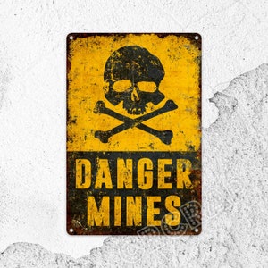 Op de afbeelding: Een rechthoekig metalen bord met een verweerde gele en zwarte afwerking. Het bovenste gedeelte bevat een zwarte afbeelding van een schedel met gekruiste beenderen. Onderaan staat de zwarte tekst DANGER MINES in hoofdletters.