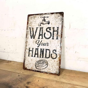 May include: A rectangular metal sign with a distressed white finish and rust-coloured edges. The sign features a black illustration of a tap at the top, the text "WASH YOUR HANDS" in a bold black font, and a drawing of a bar of soap at the bottom.