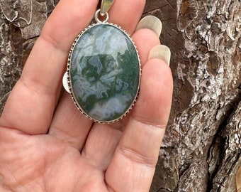 May include: Oval-shaped pendant with a silver-colored bail and setting. The pendant features a moss agate stone with green and white patterns. The stone is set in a decorative silver bezel. The pendant is held in a hand.