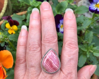 May include: A silver ring with a pink and white teardrop-shaped gemstone. The ring is set in a silver band with a rope-like design.