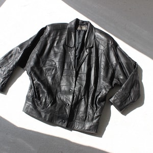 May include: A black leather jacket featuring a patchwork construction of rectangular panels, a notched lapel collar, long sleeves with dropped shoulders, and a front button closure with three visible buttons. The jacket has a structured silhouette.