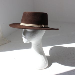 May include: A brown fedora-style hat with a flat brim and a decorative band. The hat is displayed on a white mannequin head. The hat's brim measures approximately 33 cm in diameter.