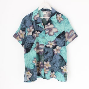 May include: Short-sleeved shirt with a floral design. The shirt has a teal base colour, with large, stylized flowers in shades of purple, white, and blue. It is hanging on a white hanger against a white background.