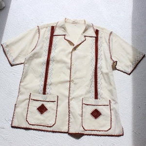 May include: A short-sleeved cream-colored button-down shirt featuring dark red decorative stitching along the collar, sleeves, and hem. Two front pockets are adorned with dark red diamond-shaped embroidery and vertical dark red stitched lines.