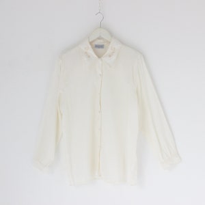 Puede incluir: Una camisa blanca de manga larga con botones y un cuello bordado con flores.