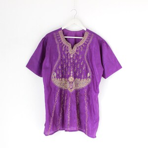 May include: A short-sleeved purple tunic with gold embroidery around the neckline and down the front. The tunic is hanging on a white hanger against a white background. The embroidery features floral and geometric patterns.