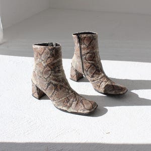 May include: A pair of ankle boots with a snakeskin pattern in shades of brown and tan. The boots have a square toe, a block heel, and a zipper closure on the side. The heels are approximately 3 inches high.