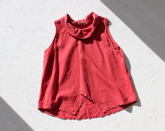 May include: A sleeveless, coral-coloured top with a cowl neck and asymmetrical hem. The top is made of a lightweight fabric, possibly linen. The garment is displayed on a white textured surface.