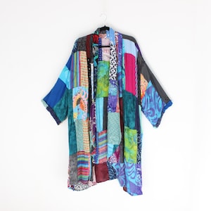 May include: A patchwork kimono in shades of blue, green, purple, and red hangs against a white background. The open-front garment features a variety of patterned fabric squares sewn together. The sleeves are wide and the overall style is bohemian.