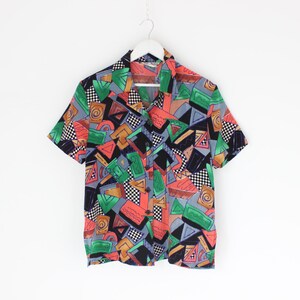 May include: Short-sleeved shirt with a vibrant geometric design. The pattern includes triangles, squares, and other shapes in red, green, black, and orange on a grey backdrop. The shirt is suspended from a white hanger.