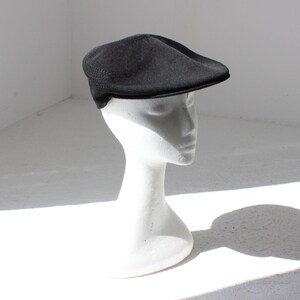 May include: A black newsboy cap on a white mannequin head. The cap features a rounded crown and a small, rigid brim. The cap is made of a textured material and is set against a white backdrop.