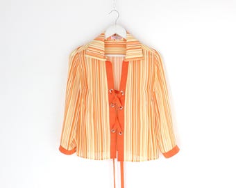 May include: Orange and cream striped blouse with a lace-up front detail. The long-sleeved shirt has a collared neckline and orange trim on the cuffs and front. The blouse is hanging on a white hanger against a white background.
