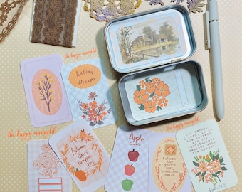 May include: A metal tin containing a landscape print and floral illustrations, surrounded by several rectangular cards featuring autumn-themed graphics, text, and floral motifs, alongside a light gray pen and decorative lace and leaf accents.