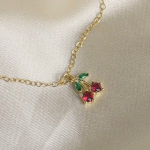 May include: A gold chain necklace with a red and green cherry charm. The charm is made of metal and has small red and green gemstones.