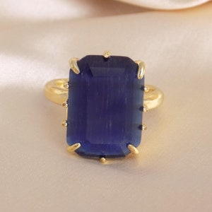 May include: A gold-toned ring featuring a large, rectangular, deep blue gemstone. The gemstone is set with four prongs and has a polished appearance. The ring is displayed on a soft, cream-colored fabric.