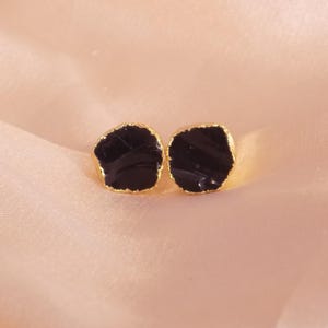 May include: A pair of black stone stud earrings with gold-colored edges. The earrings have an irregular, organic shape and are set against a soft, peach-colored background. These earrings are a stylish accessory.