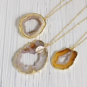 May include: Three gold-plated necklaces with natural geode pendants. The pendants feature a variety of colors, including beige, orange, and white, with gold edging. The necklaces are displayed on a white wooden surface.