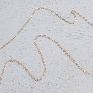 May include: A gold chain necklace with a delicate, linked design. The chain is laying on a white wooden surface.