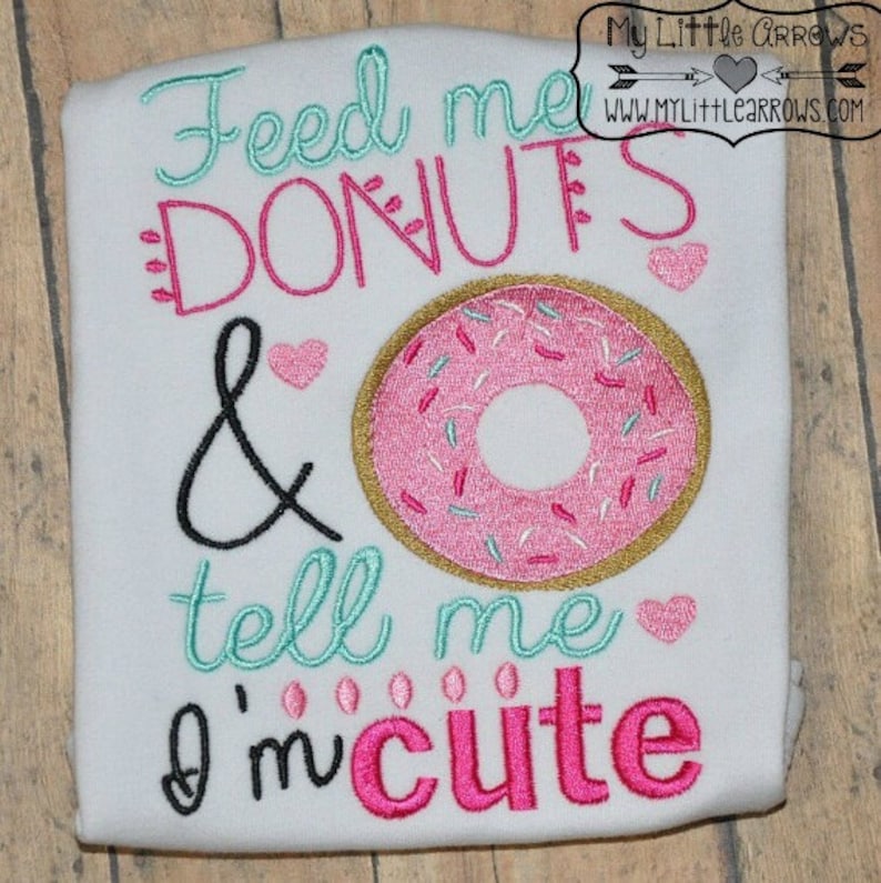 May include: White embroidered t-shirt with the text "Feed me donuts & tell me I'm cute" in pink, teal, and black. A pink donut with gold sprinkles is embroidered in the center.