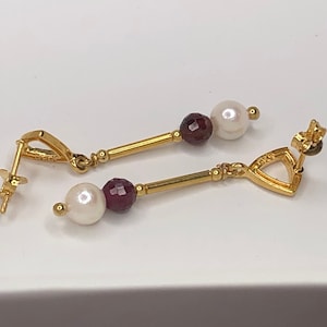 May include: A pair of gold earrings with a white pearl and a dark red gemstone. The earrings have a triangular dangle with a gold hook.