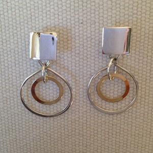 May include: Silver dangle earrings with a square top and two silver circles, one inside the other.
