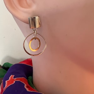 May include: Silver and gold geometric earrings with a square top and two circles hanging from it.