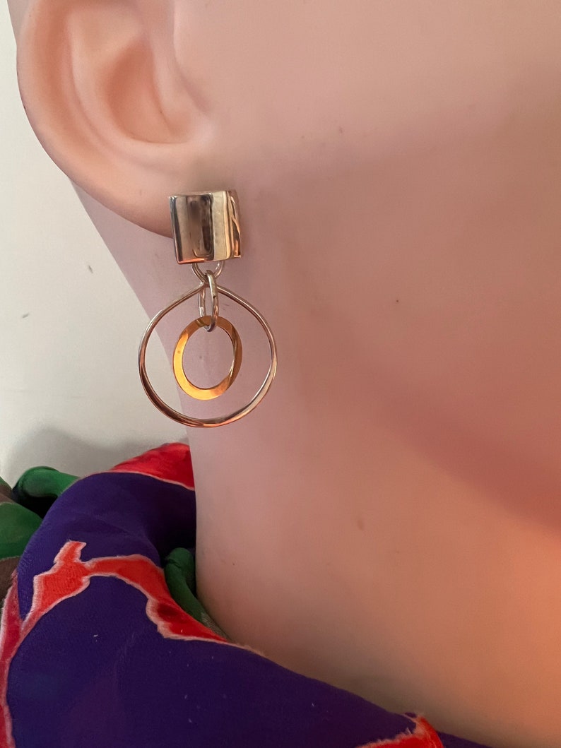 May include: Silver and gold geometric earrings with a square top and two circles hanging from it.