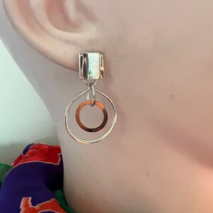 May include: Silver dangle earrings with a square-shaped top and a circle pendant. The circle pendant has a smaller circle cut out of the center.