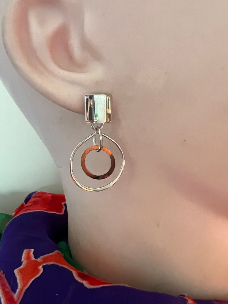 May include: Silver dangle earrings with a square-shaped top and a circle pendant. The circle pendant has a smaller circle cut out of the center.