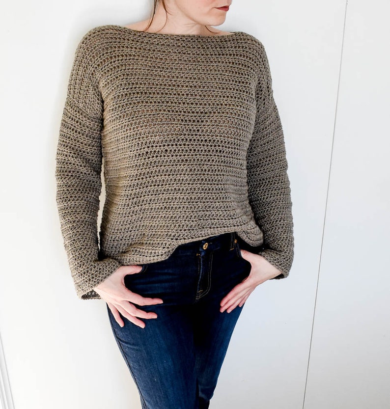 May include: A brown crocheted sweater with long sleeves and a boat neck. The sweater is worn over blue jeans.